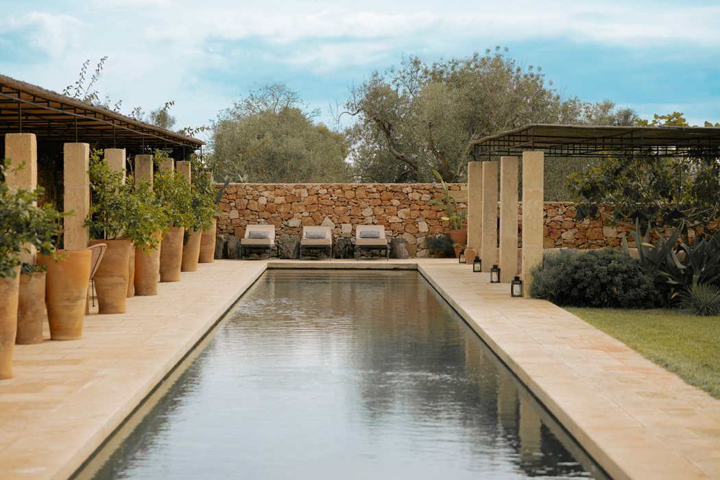 Piscina lineare circondata da pietra naturale, vegetazione mediterranea e aree relax ombreggiate.