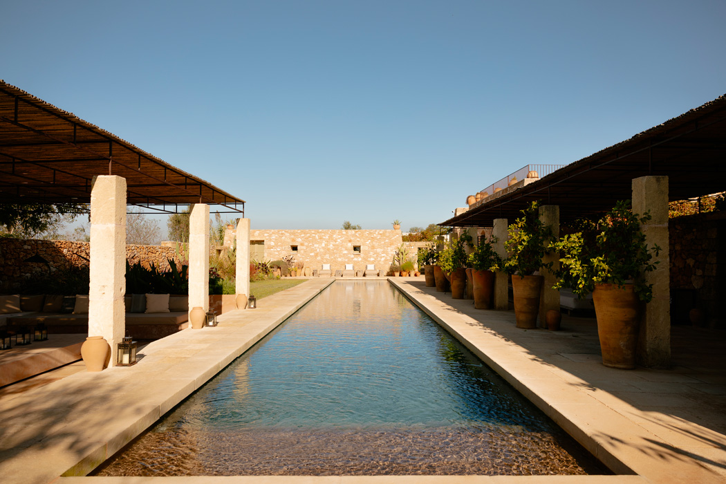 Piscina lineare circondata da pergolati, pietra naturale e vegetazione mediterranea.