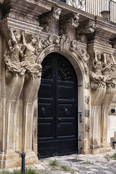 Portale barocco in pietra leccese con decorazioni scolpite e grande portone nero nel centro storico di Lecce.
