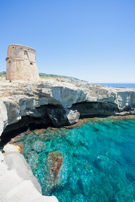 Scogliera calcarea sul mare cristallino con antica torre costiera affacciata sull’acqua turchese.