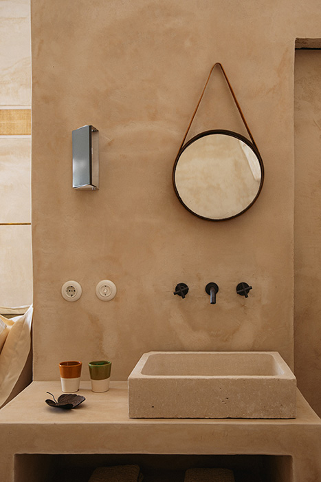 Dettaglio del lavabo in pietra con specchio circolare e pareti materiche.